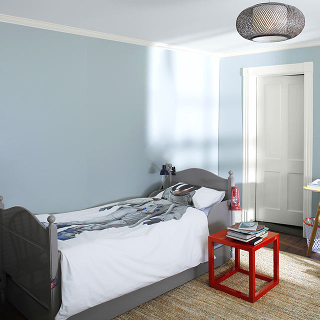 A gray-blue bedroom with white ceiling, cluttered bulletin board, bed with astronaut blanket, and red accent table.