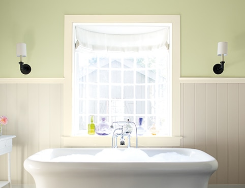Salle de bains avec lambris d’appui blanc cassé, haut des murs vert et baignoire blanche devant une fenêtre panoramique.