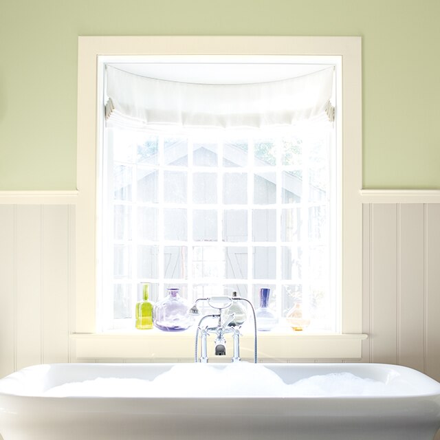 A bathroom with off-white painted wainscoting, green upper walls, and a white bathtub beneath a picture window.