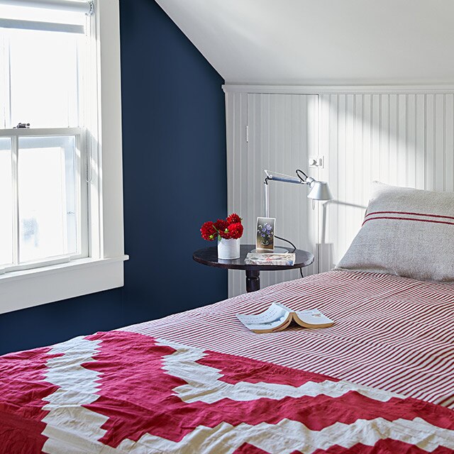 A bedroom with white-painted wainscoting, vaulted ceiling and trim, a dark blue accent wall, wood ceiling beam, and red and white bedding.