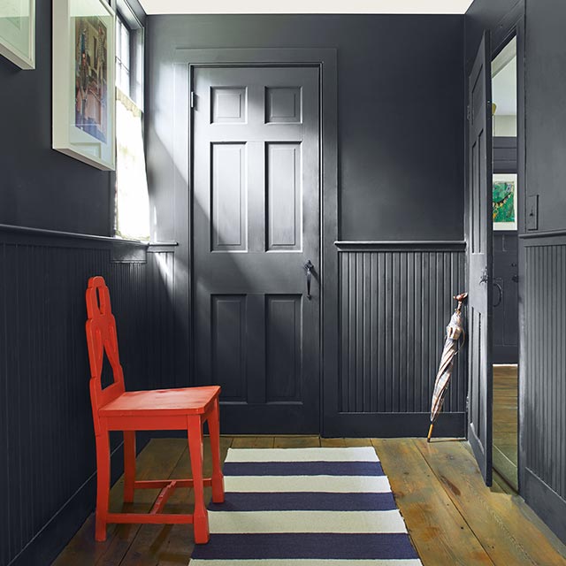 A hallway with dark gray painted walls, wainscoting and door, a bright red chair, and a blue and white striped rug.