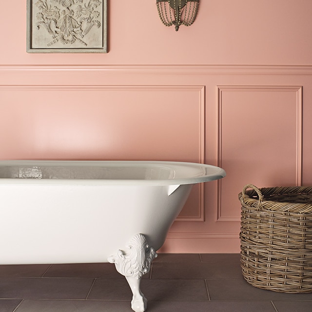 A bathroom with a pink-painted wall and wainscoting, a white clawfoot tub, a wall sconce and a basket.