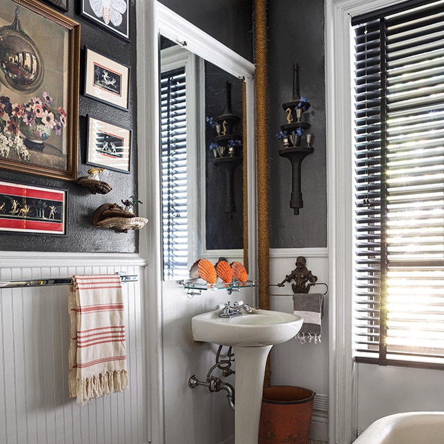 A bathroom with dark gray-painted walls, white wainscotting, trim and ceiling, a pedestal sink and eclectic artwork.