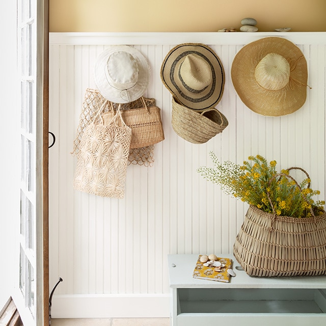 An entryway with a yellow-painted upper wall, white wainscotting, and white trim on an open door, hanging straw hats, and a gray bench.