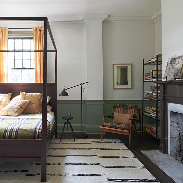 A bedroom with white-painted upper walls, dark green wainscoting, a four-poster canopy bed, black and white rug, and a fireplace.