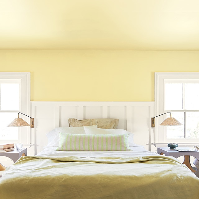A sunny bedroom with a yellow-painted wall and ceiling, white wainscotting and trim, yellow and white bedding, two reading lamps and side tables. 