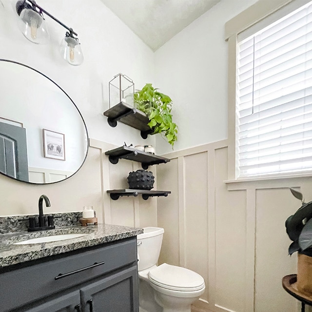 A lovely bathroom with off-white painted wainscoting and a white upper wall, wooden floors and an oval mirror over a dark-gray painted bathroom vanity with granite countertops.