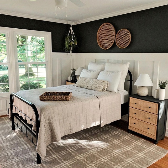 A welcoming bedroom with white-painted wainscotting and black upper walls, a wrought iron bedframe, a plaid area rug in neutral hues, and large, white-trimmed windows.