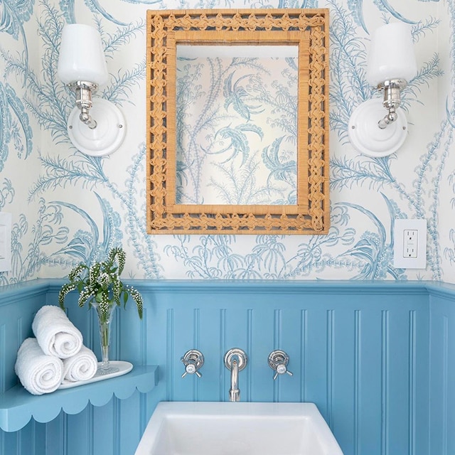 A cheery bathroom with vibrant sea blue-painted wainscoting and shelf holding towels, white and blue wallpaper, a square white sink and a playful frame highlighting the wallpaper design.