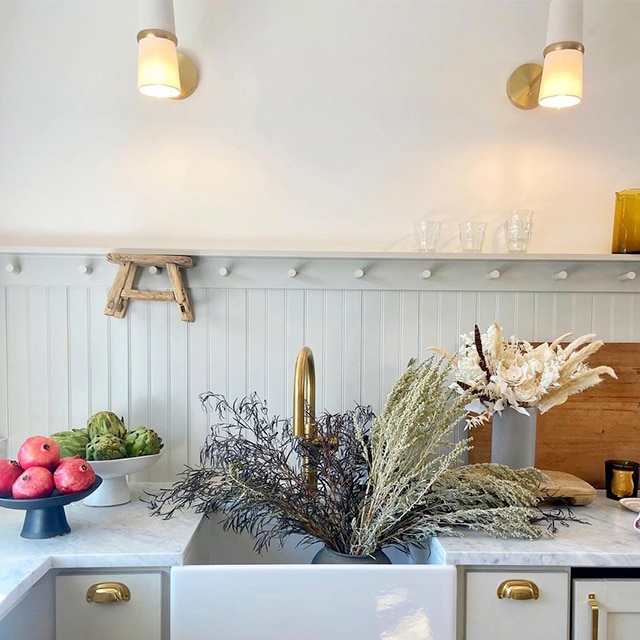 An off-white kitchen with pale green-painted wainscoting and cabinetry, white upper walls, white countertops, and a farmhouse sink filled with flowers.