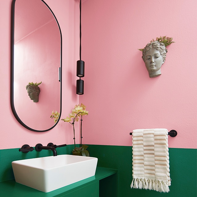 A bathroom with modern fixtures, an oval mirror and décor modeled after an ancient bust features a two-toned wall with bright pink on the upper and a deep green on the lower. 