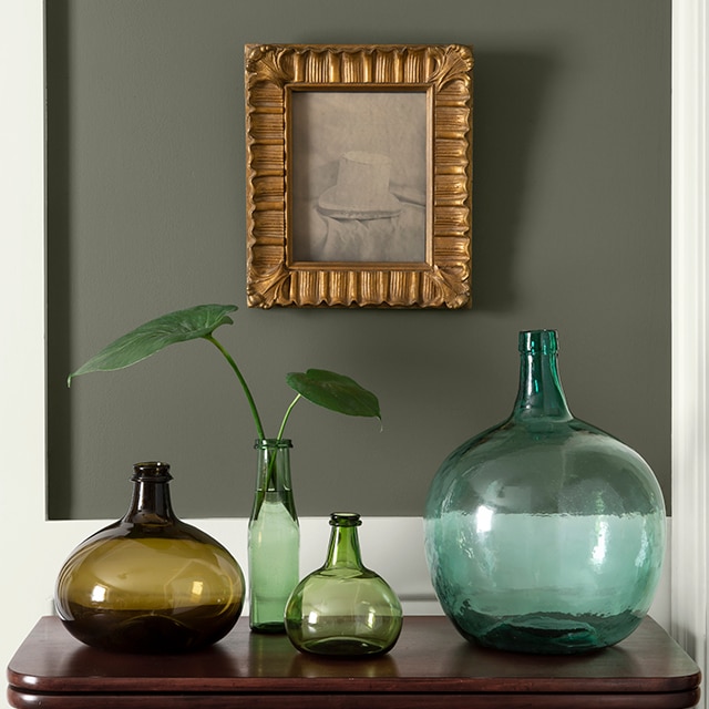 A close-up of an olive green-painted hallway and off-white wainscotting with an antique wooden table holding coloured vases beneath a small, framed painting.