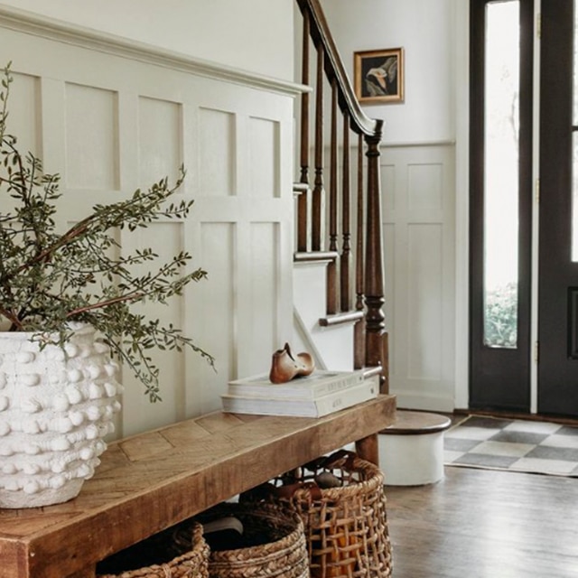 An airy off-white-painted hallway with a glass panelled front door, a staircase with wooden banister, and three large baskets beneath a narrow wooden console table holding a large white planter.