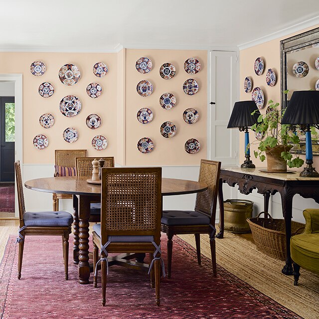 In a peachy-pink painted dining room adorned with decorative plates on the walls, a wooden table, four chairs and a side table sit atop a burgundy rug on a light hardwood floor. 