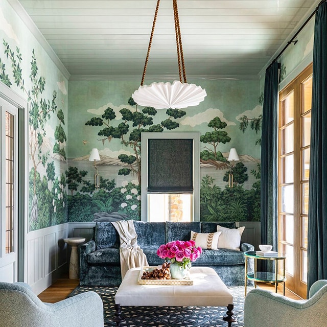 A lovely living room parlor with blue velvet couch, two armchairs, a patterned rug  and white-painted wainscoting beneath wallpaper featuring lush outdoor scenery.