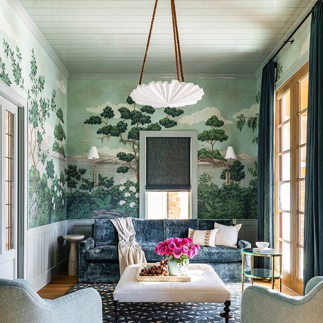 A lovely living room parlor with blue velvet couch, two armchairs, a patterned rug and white-painted wainscotting beneath wallpaper featuring lush outdoor scenery.