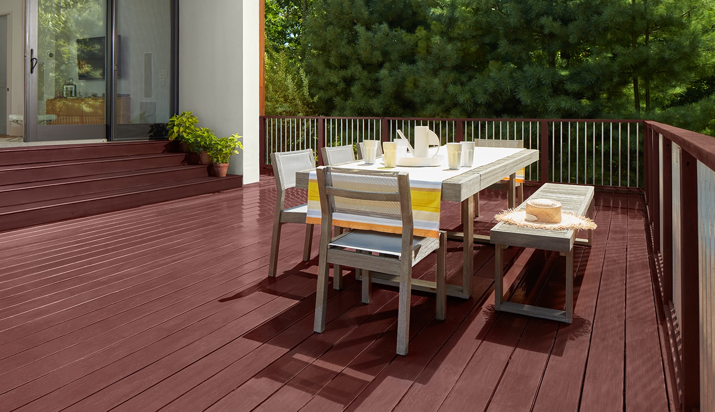 A burgundy red stained deck with outdoor dining furniture and stained steps leading to a home’s large sliding glass doors.