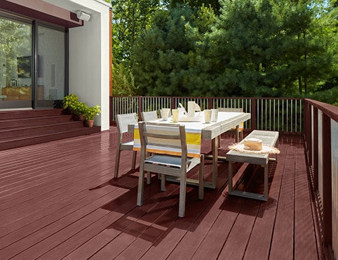 A burgundy red stained deck with outdoor dining furniture and stained steps leading to a home’s large sliding glass doors.