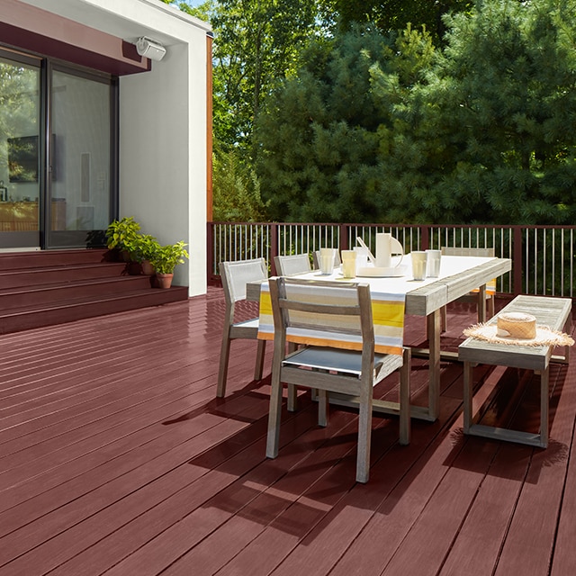 A burgundy red stained deck with outdoor dining furniture and stained steps leading to a home’s large sliding glass doors.