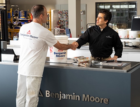 A retailer stands behind a Benjamin Moore counter, assisting a contractor in purchasing paint.