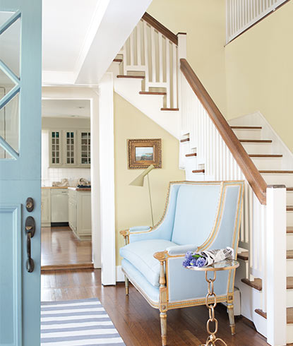 A bright entryway in neutral shades includes a staircase, blue chaise and light blue front door.