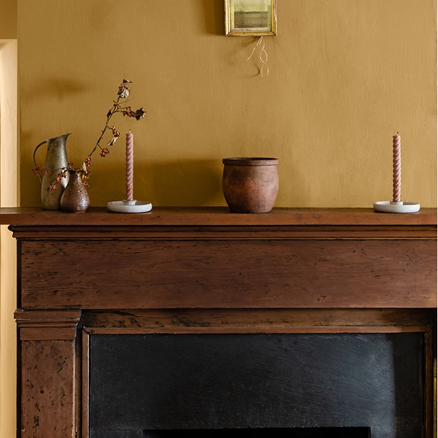 A rich, gold-brown painted wall with a small piece of framed art, and dark wood fireplace mantel with  rustic pottery and candles.