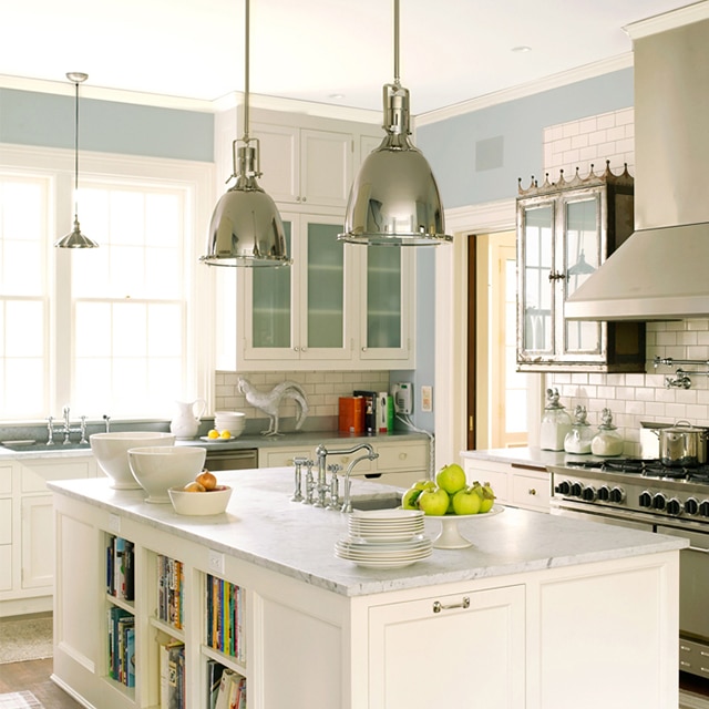 A beautiful open kitchen with light-blue painted walls, white cabinets and an island with books shelves and a sink, a white tile backsplash with antique wall cabinets, and pendant lights.