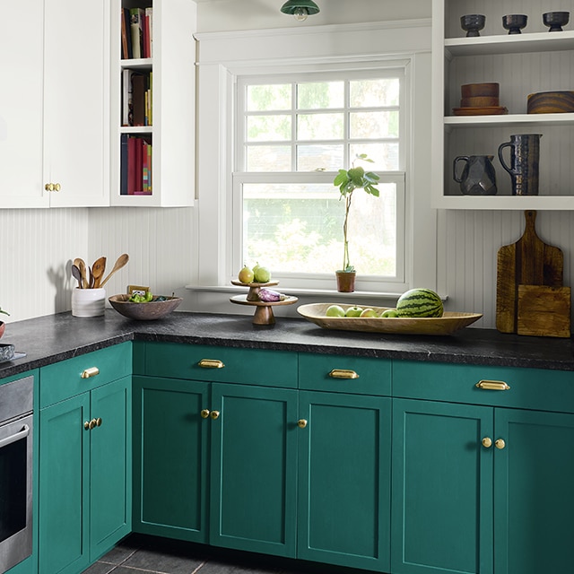 A kitchen corner with beautifully contrasting white painted walls, shelves and upper cabinets, and dark green-painted lower cabinets with gold hardware, and a black counter top.