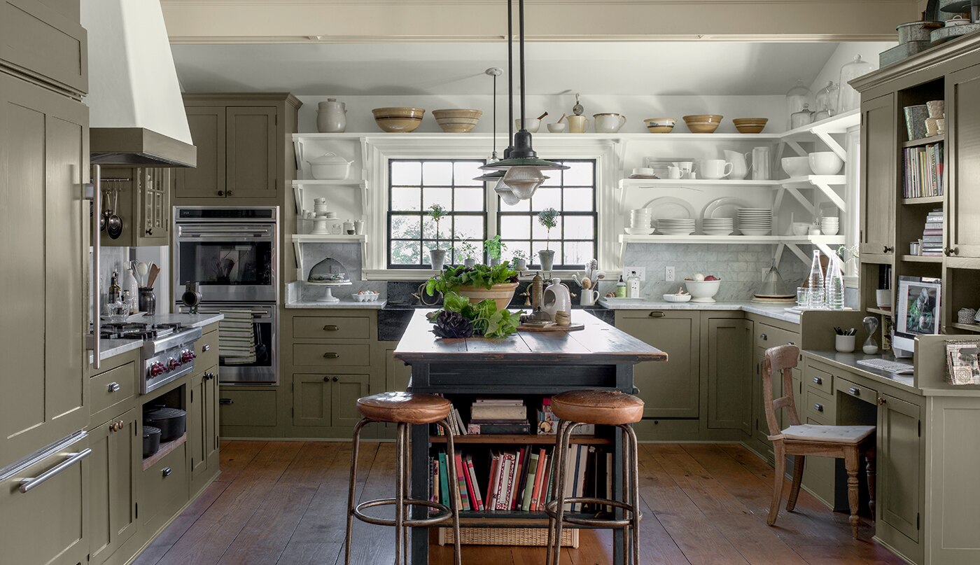 An inviting, open kitchen with sage green-painted cabinets, white open shelving with a collection of white stoneware, a rustic wooden center island and wood floor.