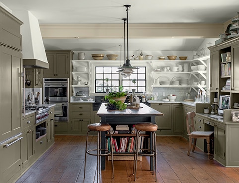 An inviting, open kitchen with sage green-painted cabinets, white open shelving with a collection of white stoneware, a rustic wooden centre island and wood floor.