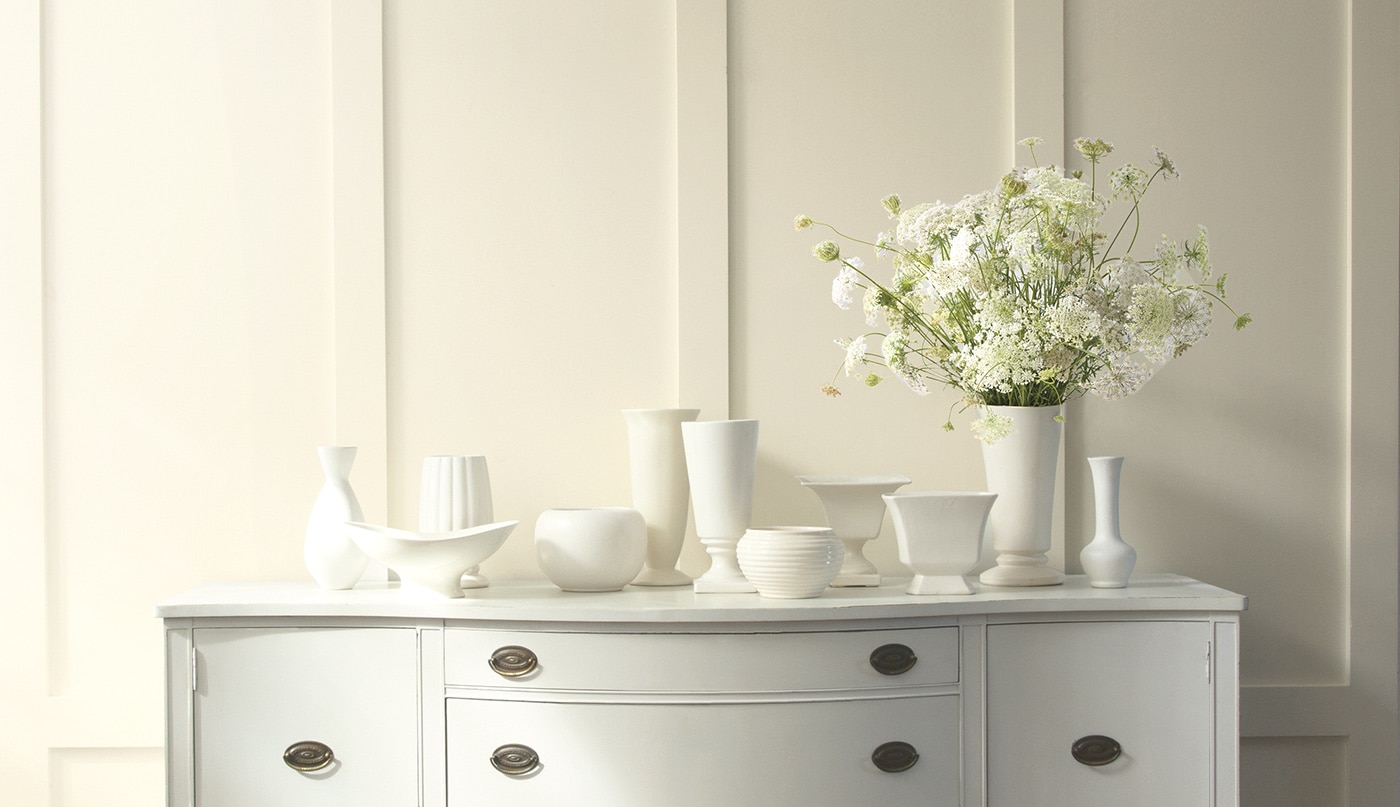 An off-white wall with floor-to-ceiling wainscoting and multiple white vases placed on top of a white dresser.