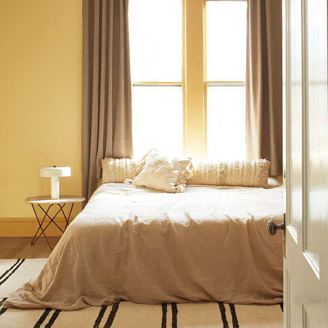 Chambre à coucher apaisante au mur jaune souci, moulures dorées, plafond blanc, ventilateur de plafond noir, literie beige, tapis rayé et porte beige.