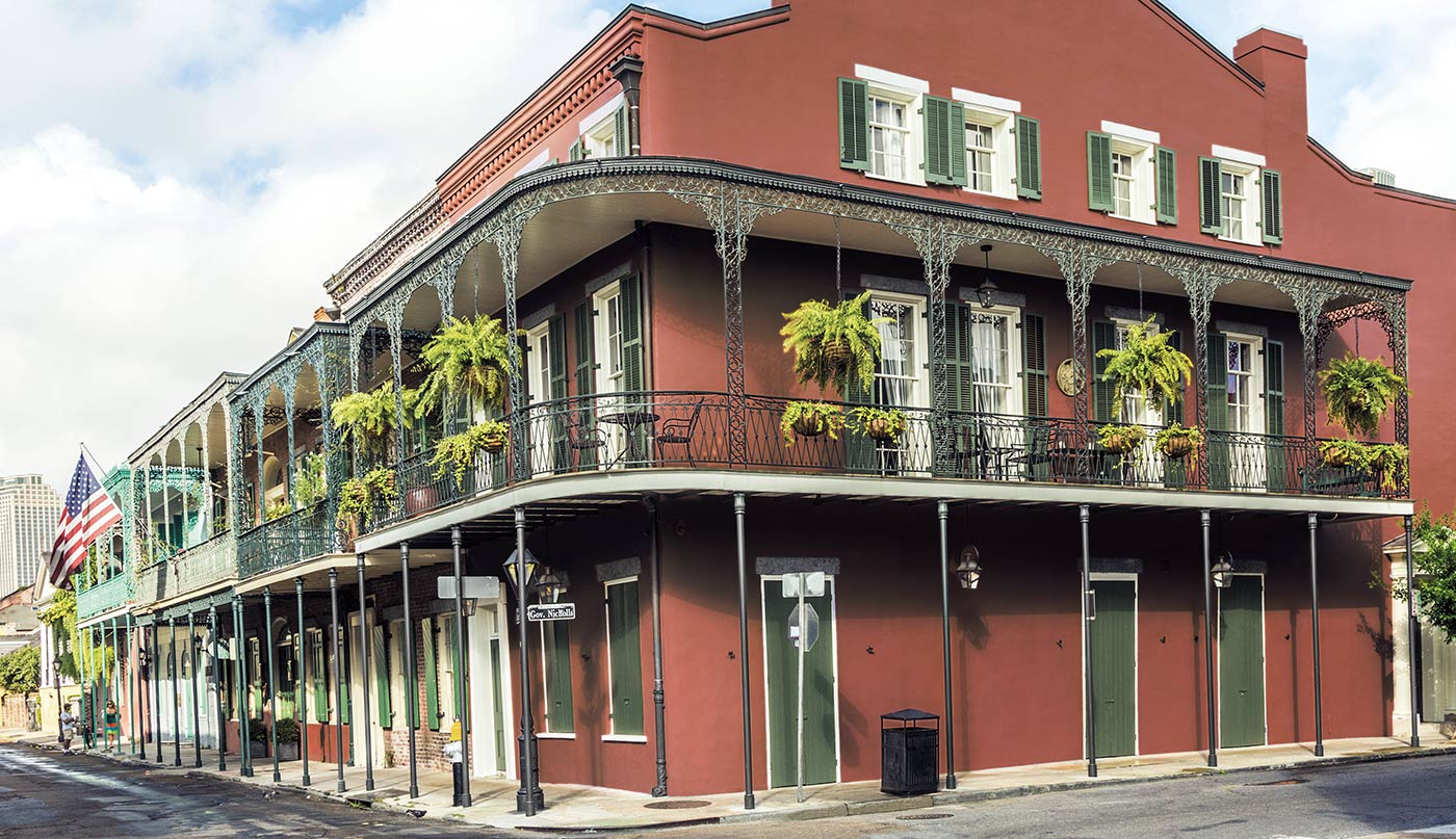 A beautiful, historic 3-story reddish brown-painted corner building with green shutters, a wrap-around wrought iron balcony, and hanging plants.