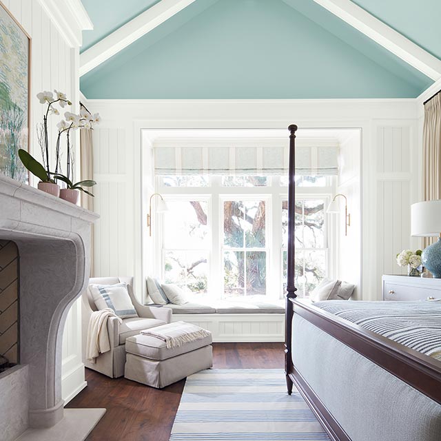 A sunlit bedroom with white-painted shiplap walls, trim and ceiling beams, a blue-painted upper wall and vaulted ceiling, blue and white decor, and a fireplace.