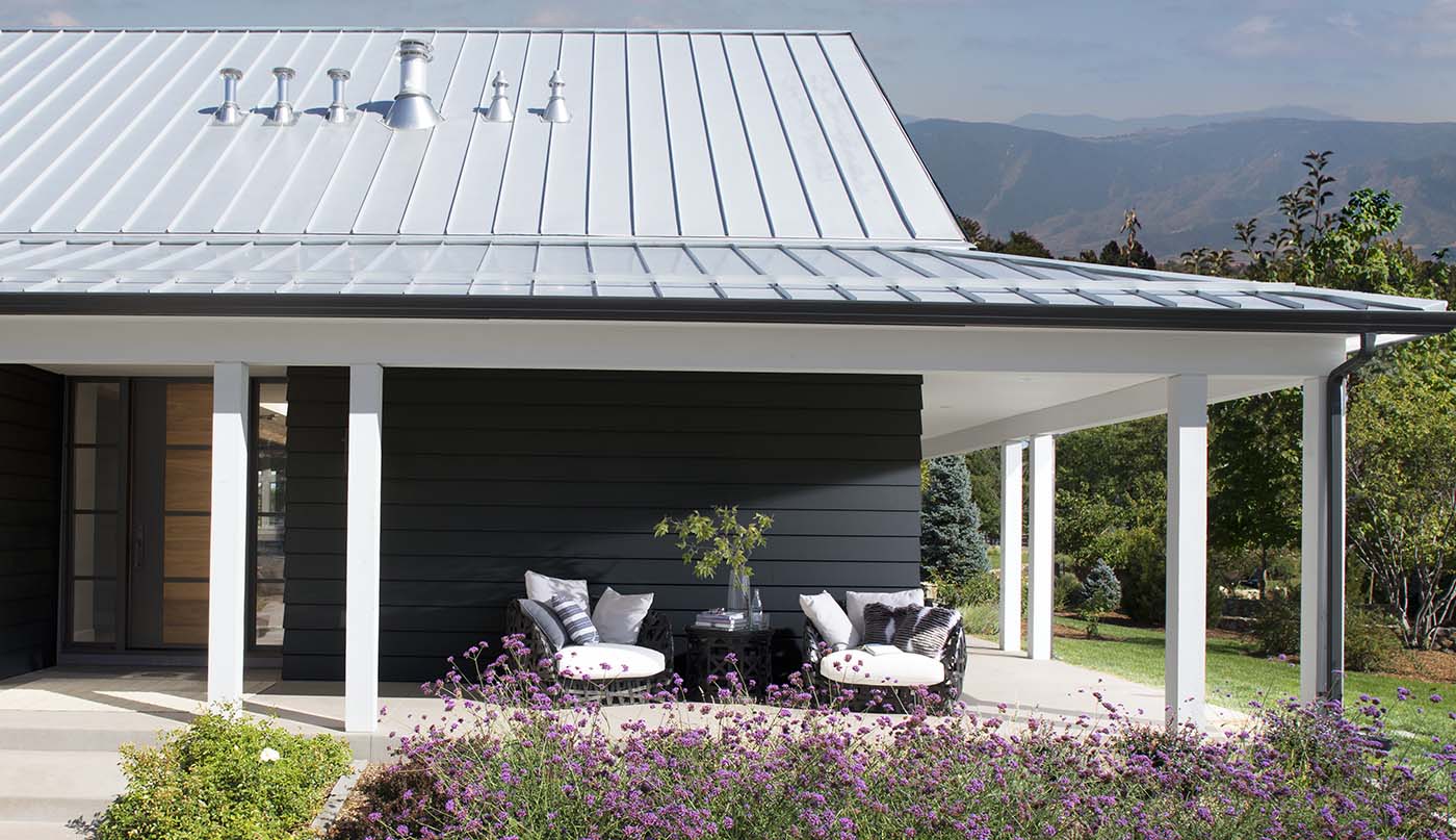 Una casa con revestimientos exteriores negros, un acogedor patio envolvente con postes, techo y cómodos muebles blancos y flores silvestres de color púrpura en el frente.