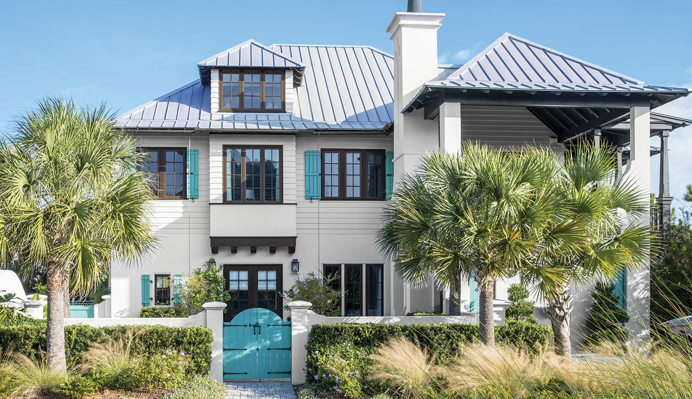 Large off white-painted home with brown trim, mahogony front doors and turquoise gate and shutters with palm trees.