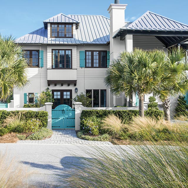Large off white-painted home with brown trim, mahogony front doors and turquoise gate and shutters with palm trees.