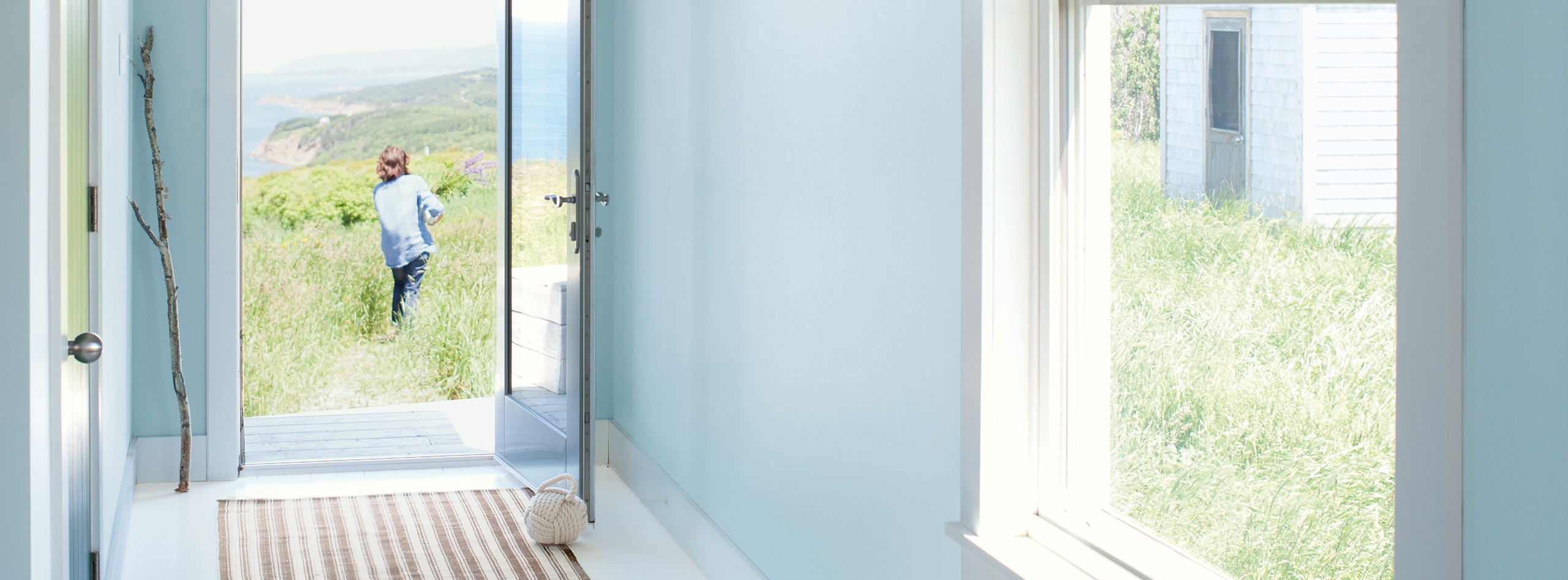 Hallway with light blue walls, white trim, and open door, framing a woman with flowers walking through a field to the ocean.