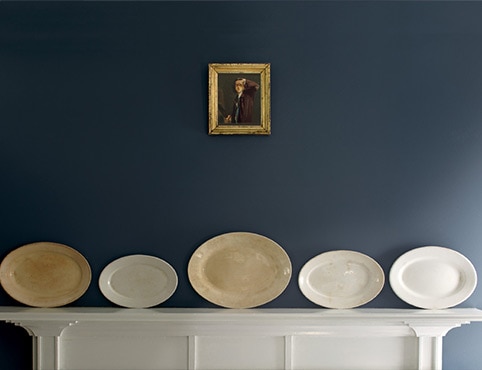 A white wood mantel features five decorative plates of varying sizes in neutral tones horizontally placed against a blue wall; a small antique painting hangs above.