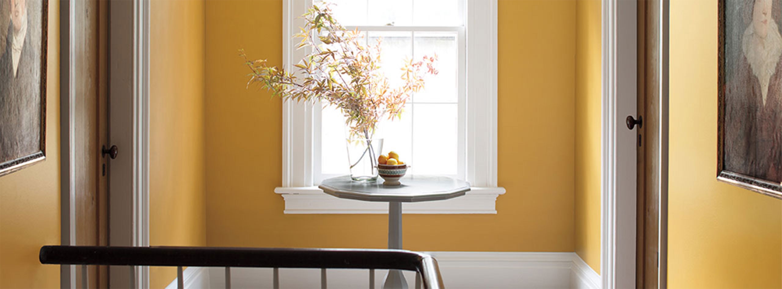 Un couloir peint en jaune arborant une table en bois et des fleurs, deux portes, une fenêtre et des moulures blanches.