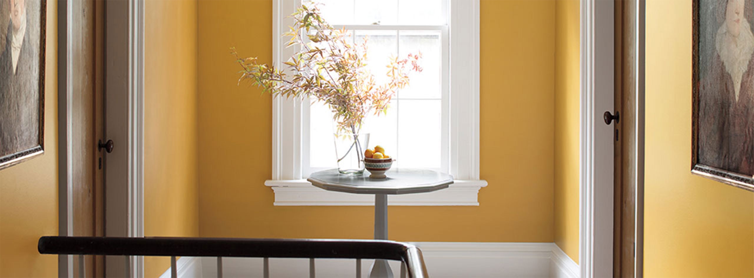 Pasillo pintado de amarillo con mesa de madera y flores, dos puertas, una ventana y marcos pintados de blanco.