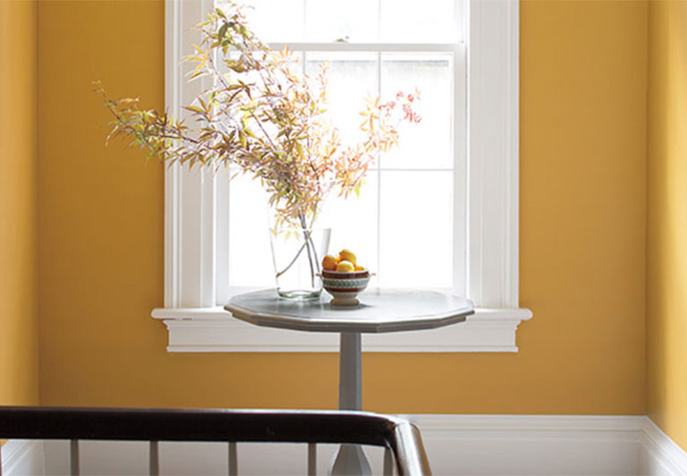 Un couloir peint en jaune arborant une table en bois et des fleurs, deux portes, une fenêtre et des moulures blanches.