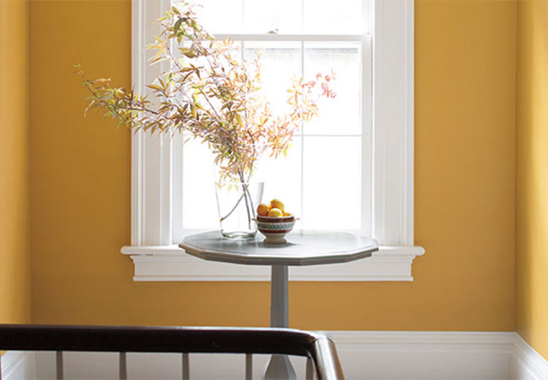 Pasillo pintado de amarillo con mesa de madera y flores, dos puertas, una ventana y marcos pintados de blanco.