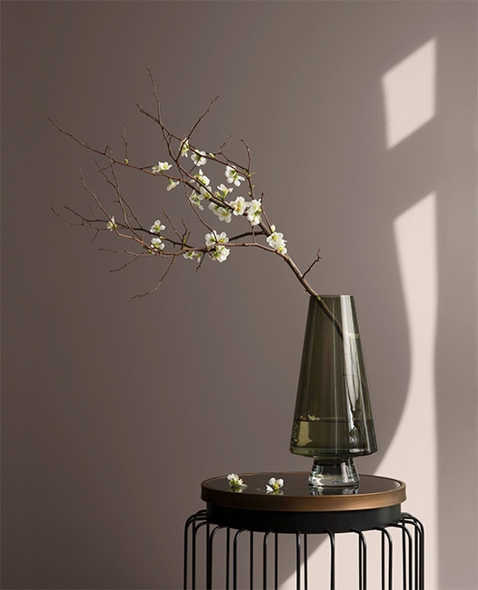 Стена, окрашенная в бежевый цвет, с современным приставным столиком и вазой с веткой белых цветов.