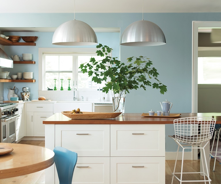 Une cuisine moderne aux murs peints en bleu pâle arborant des luminaires suspendus, des armoires blanches et un tabouret en métal.
