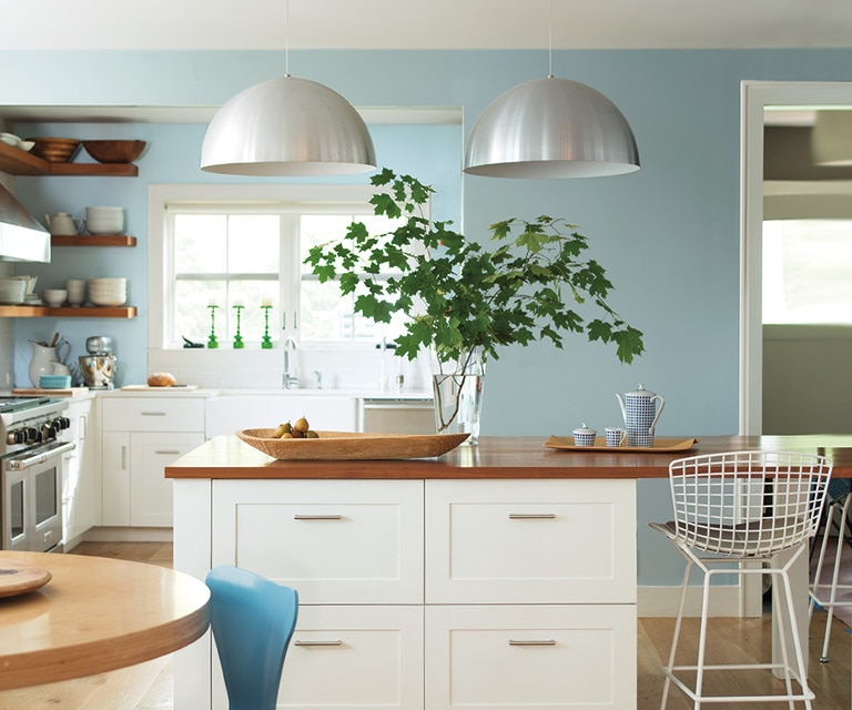 Cocina moderna con iluminación cenital, paredes pintadas de azul claro, encimeras blancas y asientos de metal.