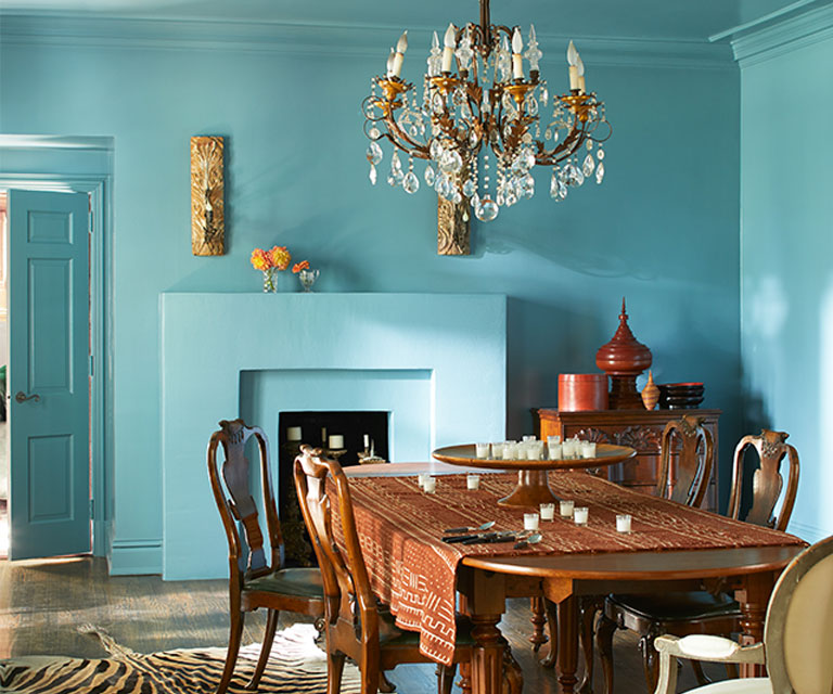 Elegante comedor con paredes, techo y chimenea pintados de azul y una lámpara de araña sobre una gran mesa de madera con sillas. El estilo es clásico y con un toque exótico.