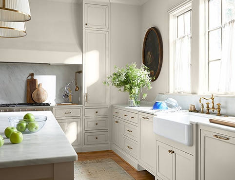 White-painted kitchen with ample counter space, farmhouse sink, kitchen island, and various plants and flowers.
