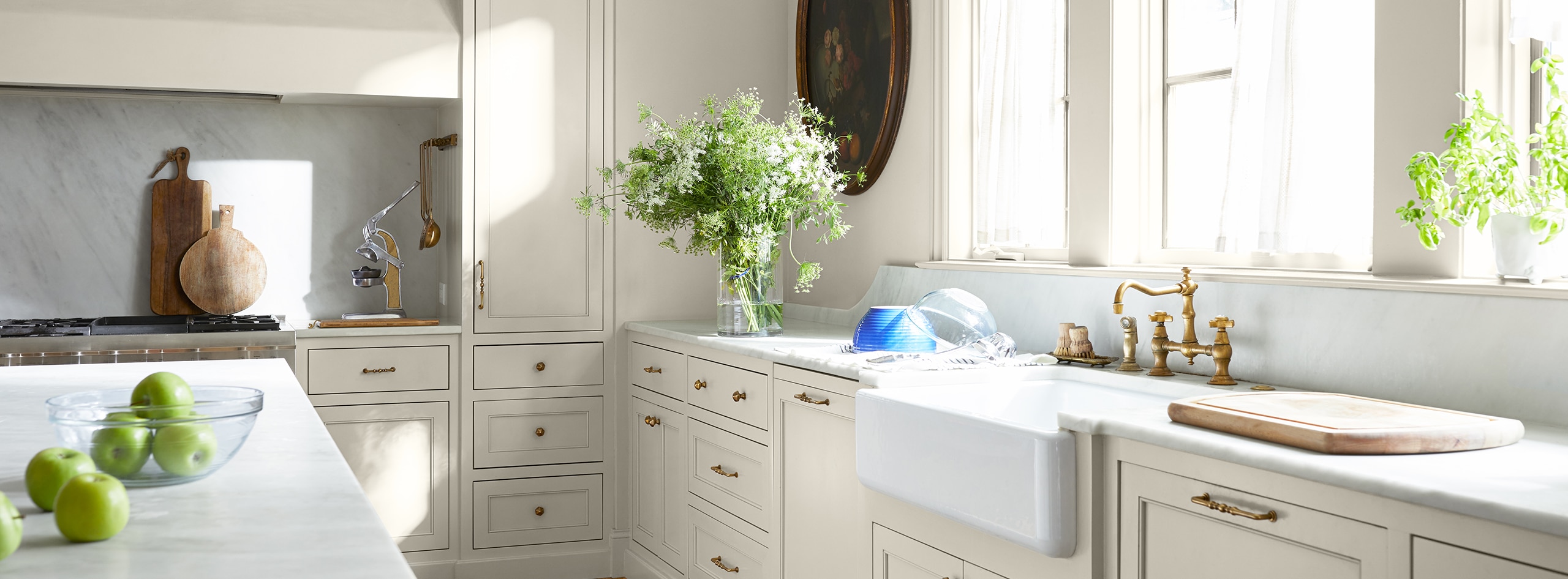 Cocina pintada de blanco con una amplia encimera, un lavabo de granja, una isla de cocina y varias plantas y flores.