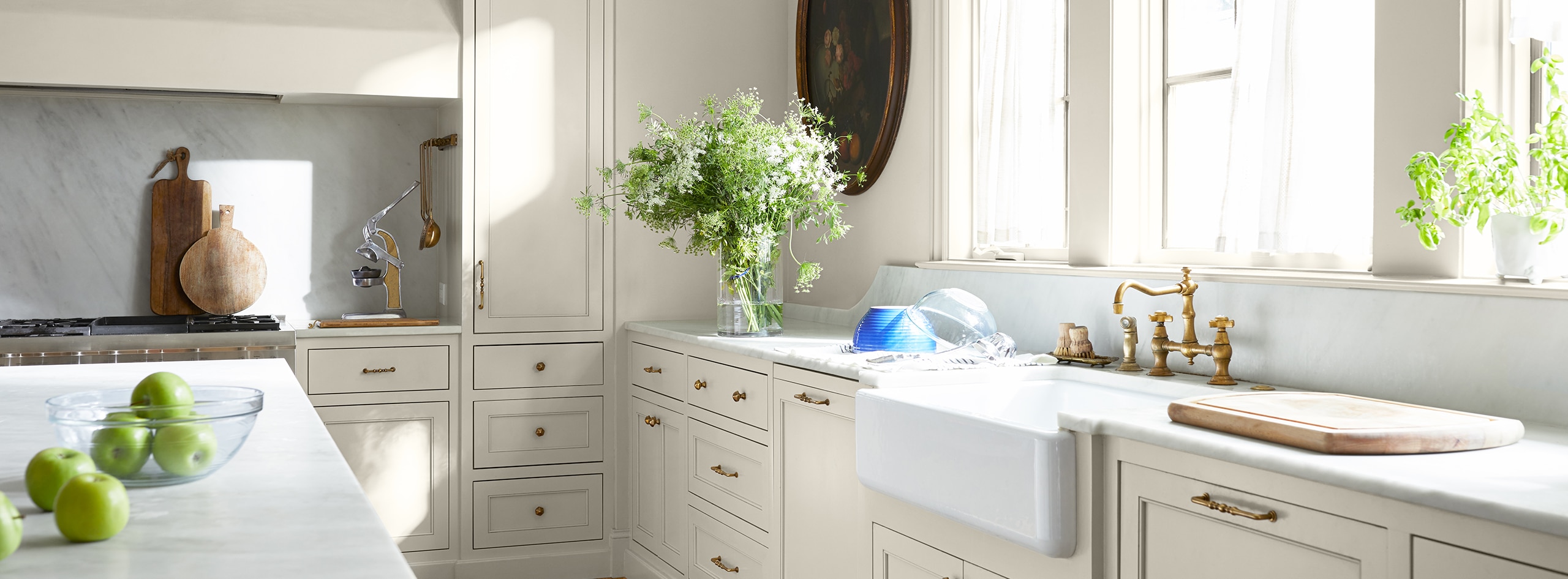 White-painted kitchen with ample counter space, farmhouse sink, kitchen island, and various plants and flowers.